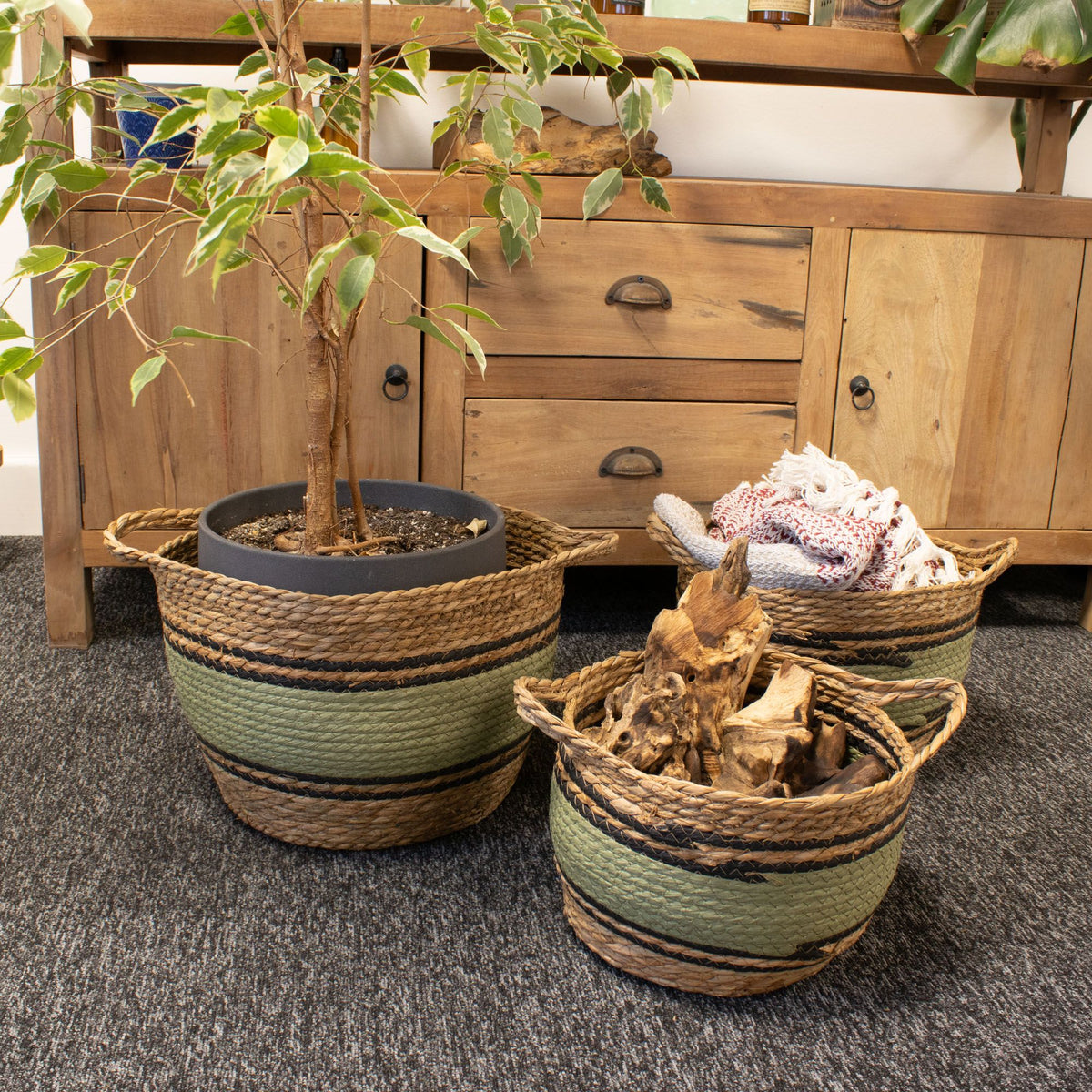Three black and green baskets holding a potted plant, folded textiles and logs beside a wooden sideboard