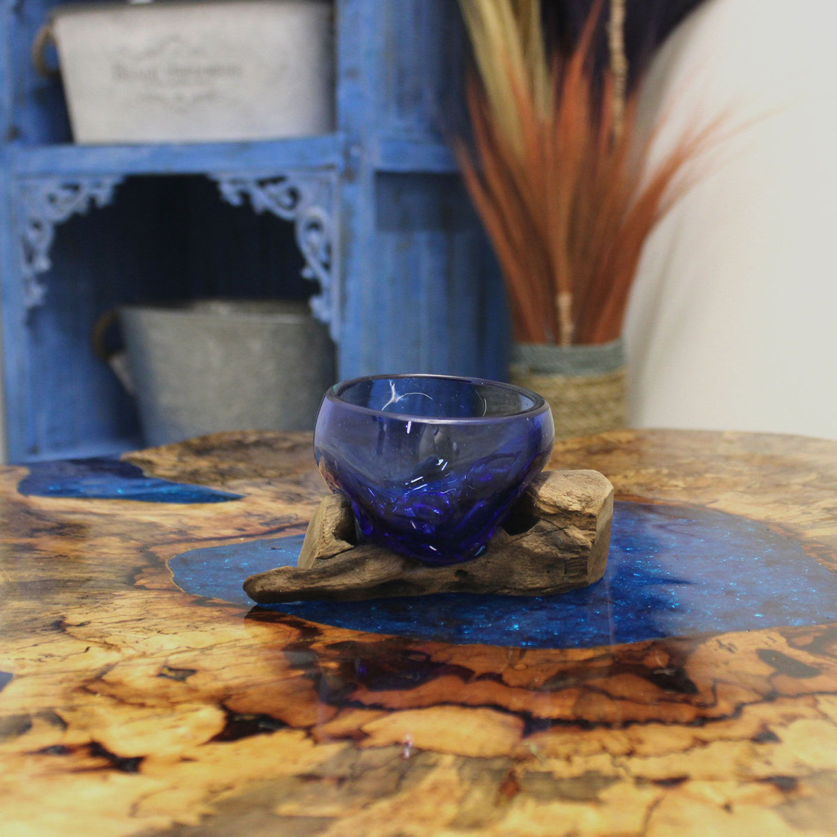 Blue glass bowl on a natural wood base, styled with a blue cabinet and dried foliage behind