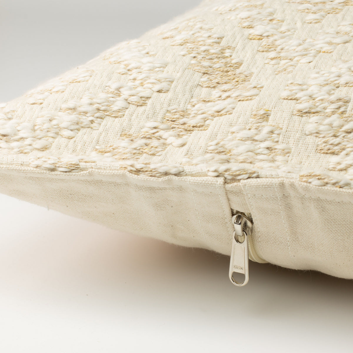 Close-up of a cream-colored textured throw pillow with a woven pattern and a silver zipper pull, shown on a light gray background.