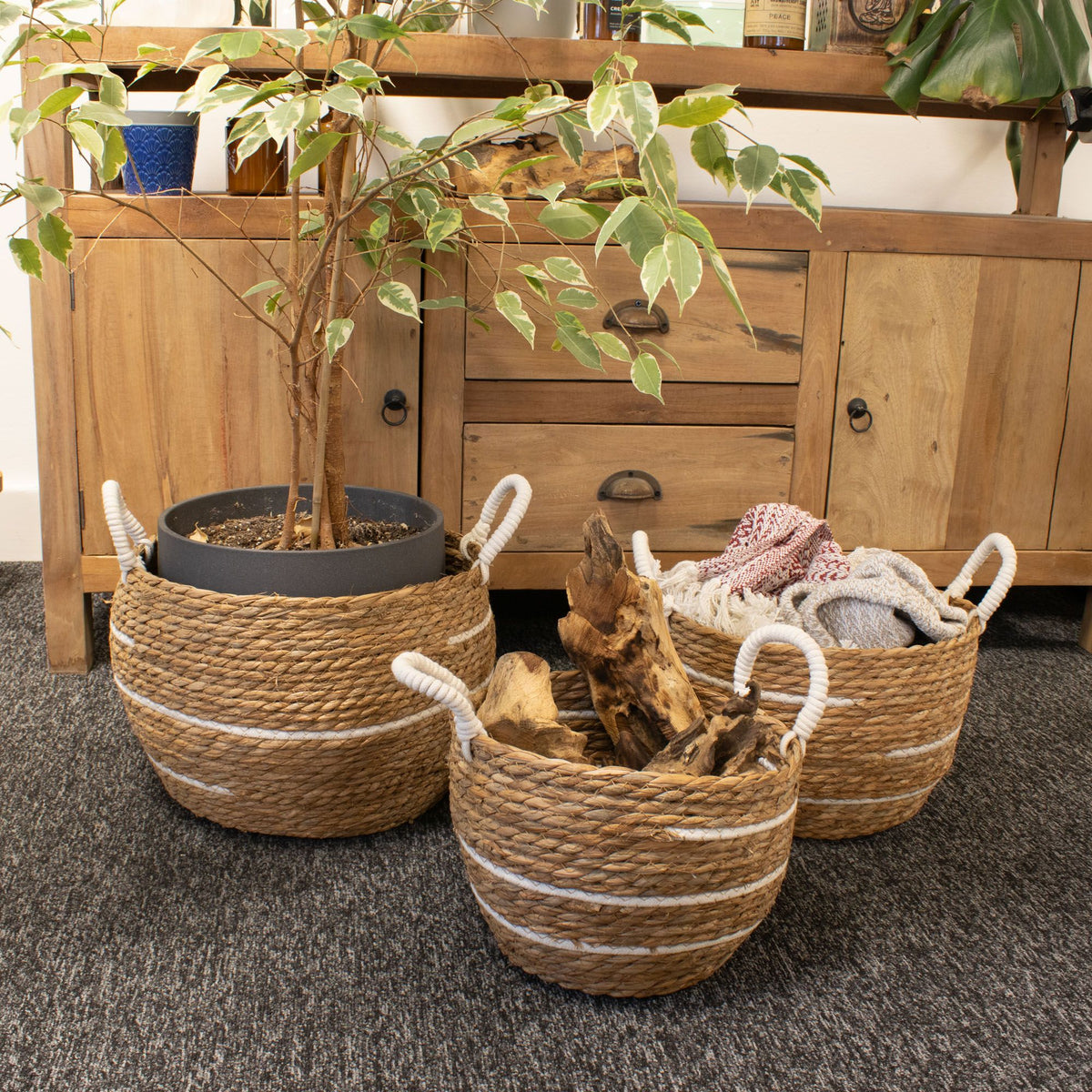 Three striped baskets holding a potted plant, folded textiles and logs beside a wooden sideboard