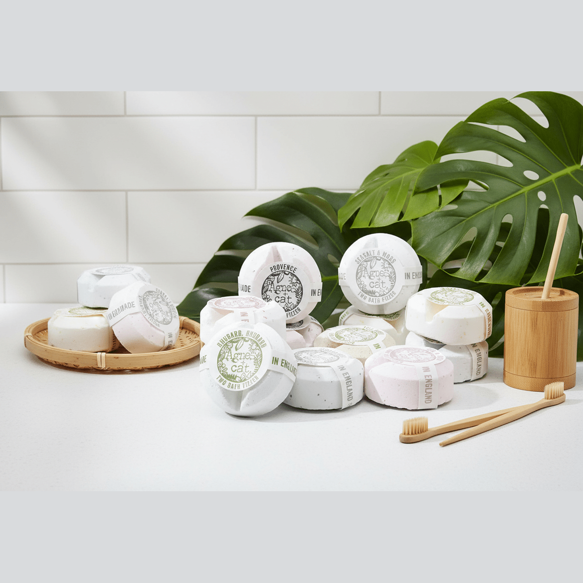 Various bath fizzers on a white surface with a bamboo basket and toothbrushes, against a white tiled wall with green leaves.