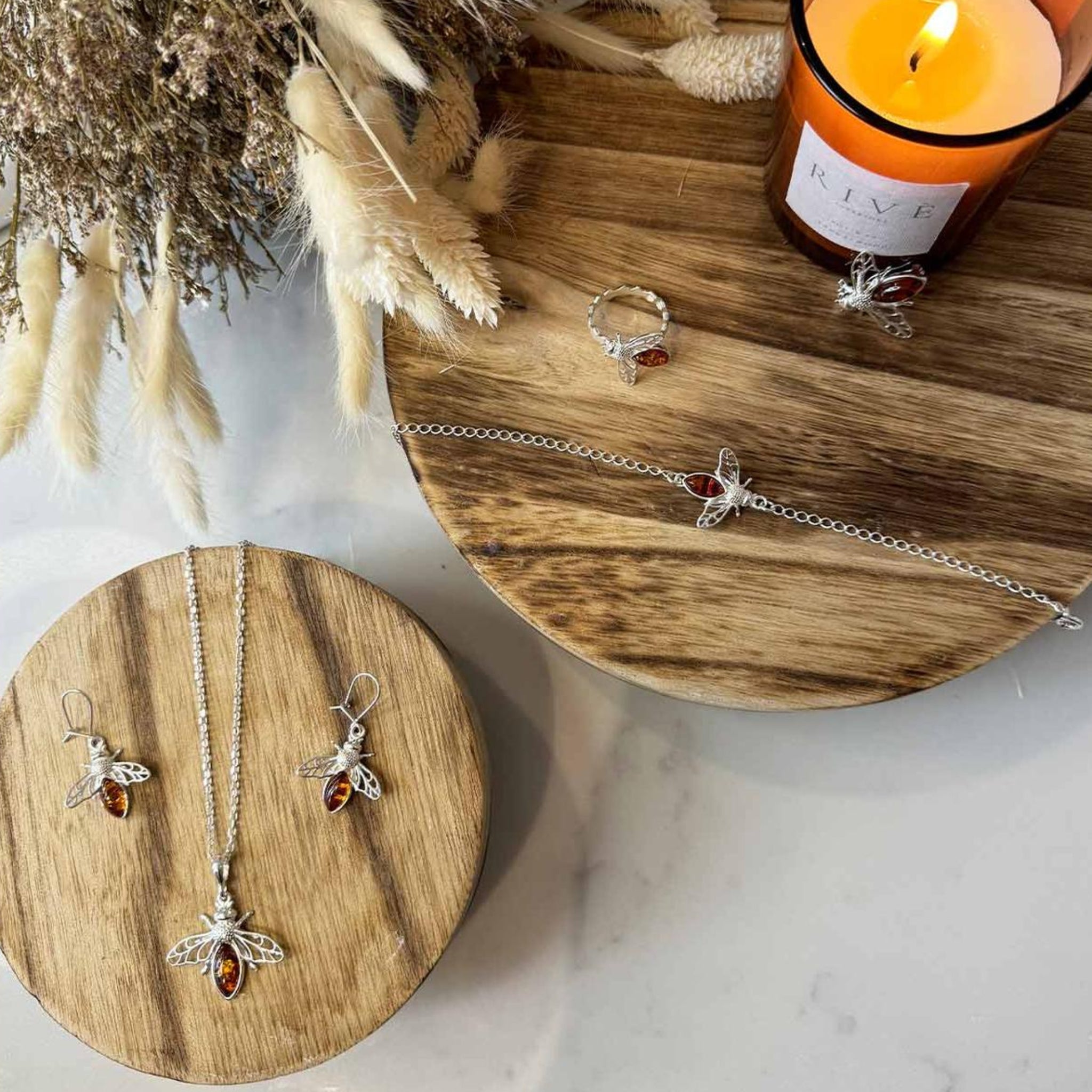 A Bee Jewellery set, including necklaces and rings, on a wooden surface with a candle in the background.