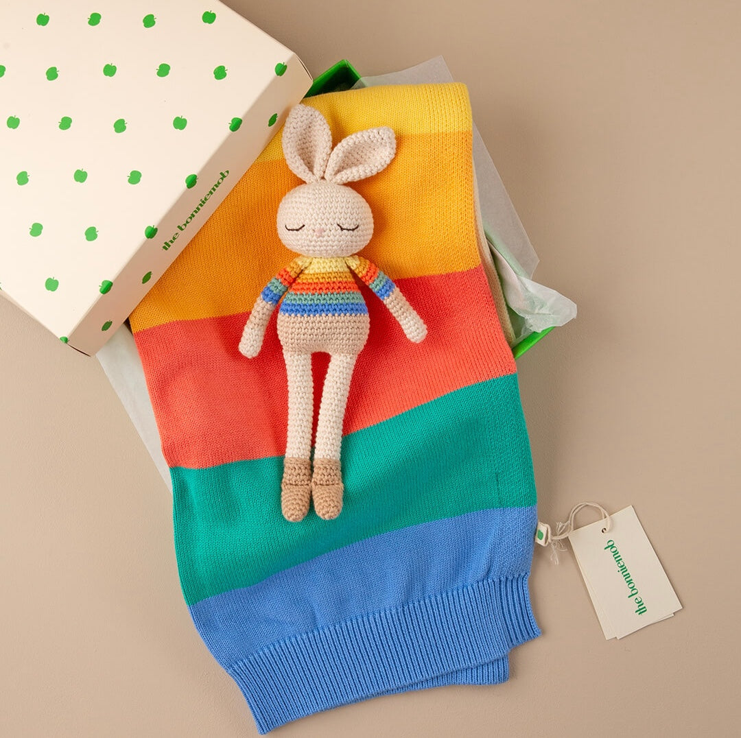 Bonnie Bunny resting on folded rainbow knitwear beside The bonniemob branded box