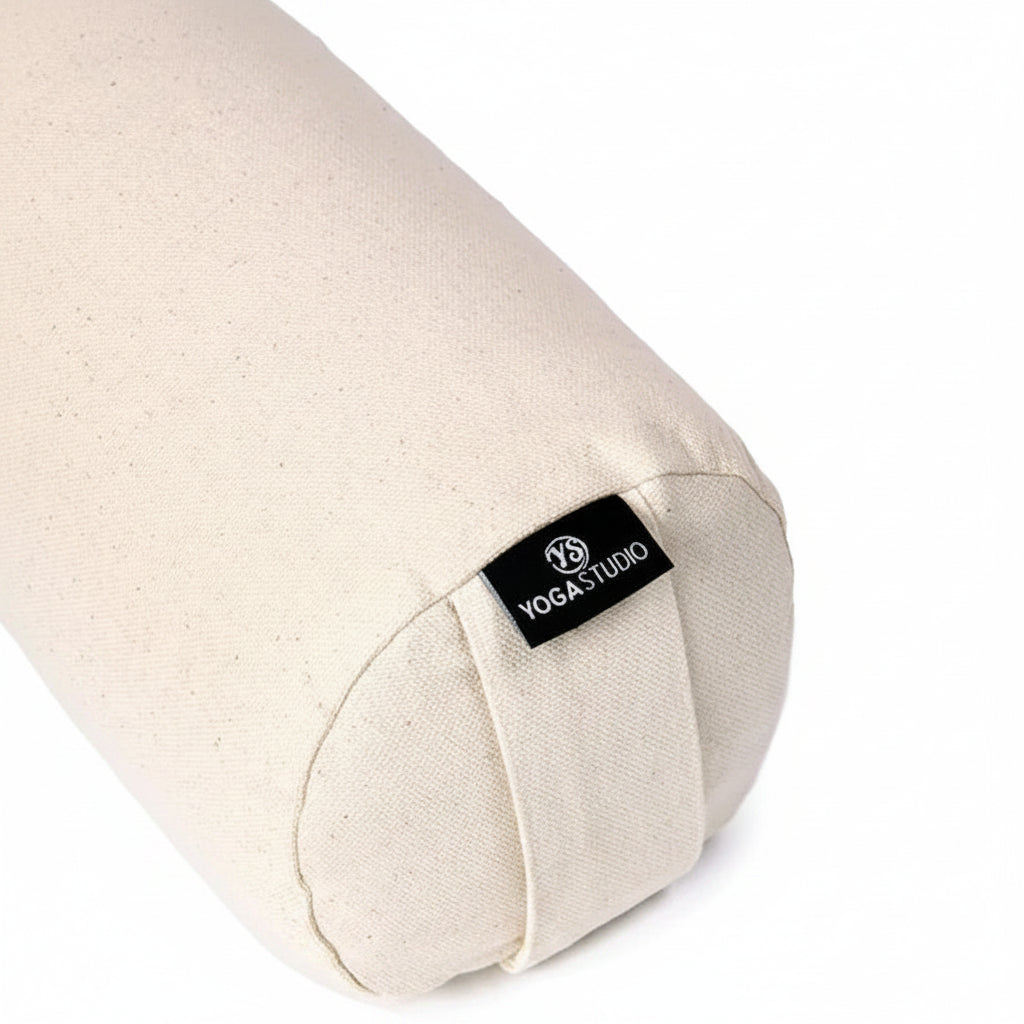 A close-up of a cylindrical yoga bolster pillow made of natural beige canvas fabric, featuring a black fabric label with the brand name 'YOGA STUDIO'.