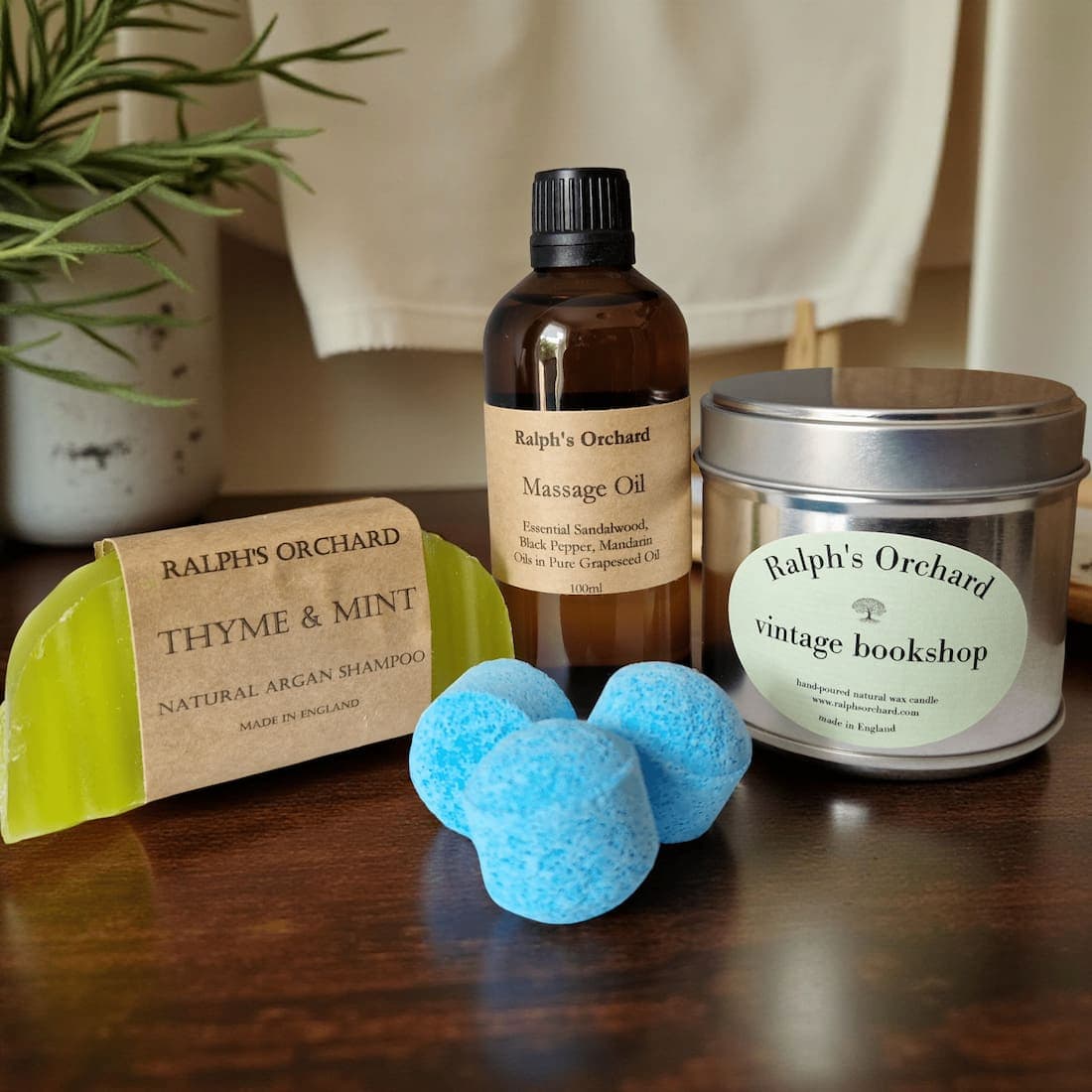 Collection of Ralph's Orchard products, including a bottle of massage oil, a candle, a shampoo bar, and bath bombs on a wooden surface.