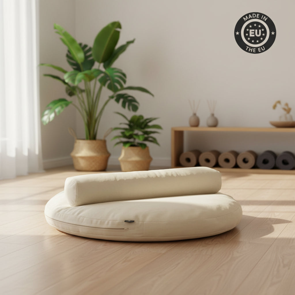 A cream-coloured floor yoga cushion set with a round base and a matching rolled lumbar pillow, placed on a light wood floor in a minimalist room with potted plants and yoga mats in the background. A 'MADE IN THE EU' badge is visible in the top right corner.