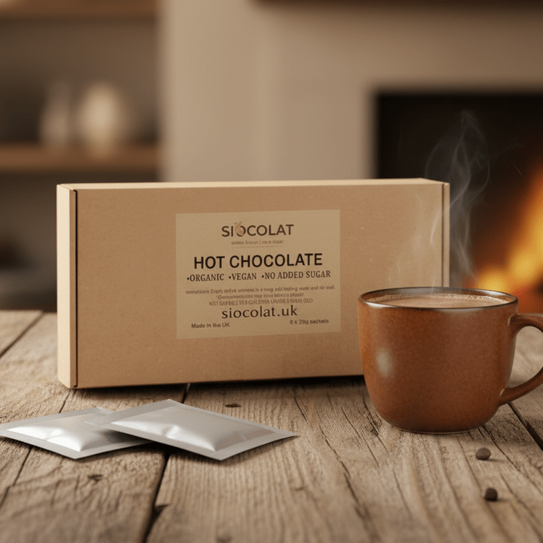 Box of siocolat hot chocolate with a steaming mug on a wooden table in front of a fireplace.