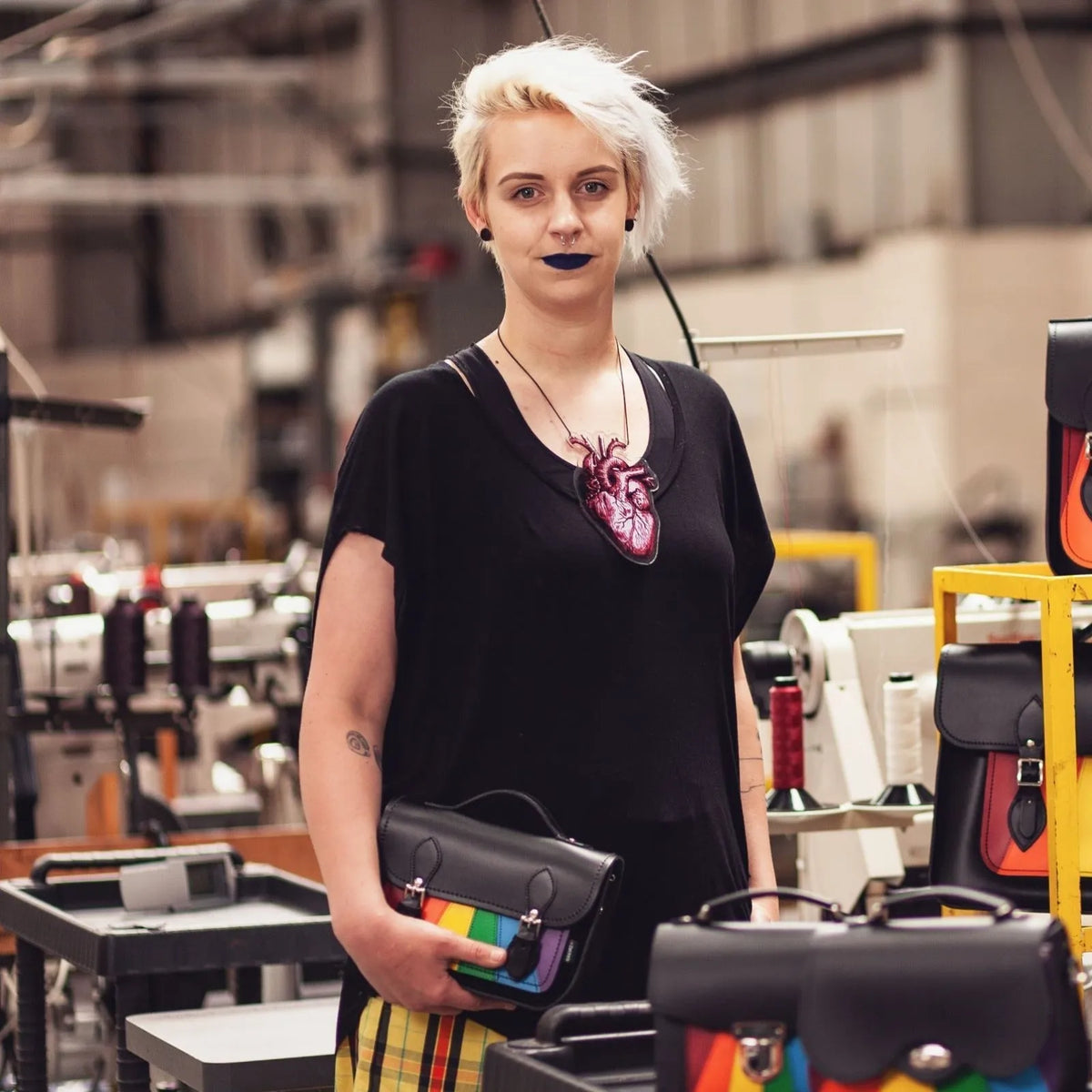 Model holding the Zatchels Pride rainbow midi satchel in a workshop, with other bag styles nearby.