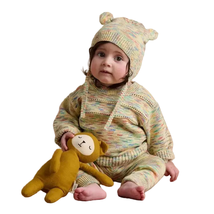 A baby wearing a knitted organic cotton jumper and matching beanie hat with bear ears, sitting and holding a yellow plush monkey toy against a plain white background.