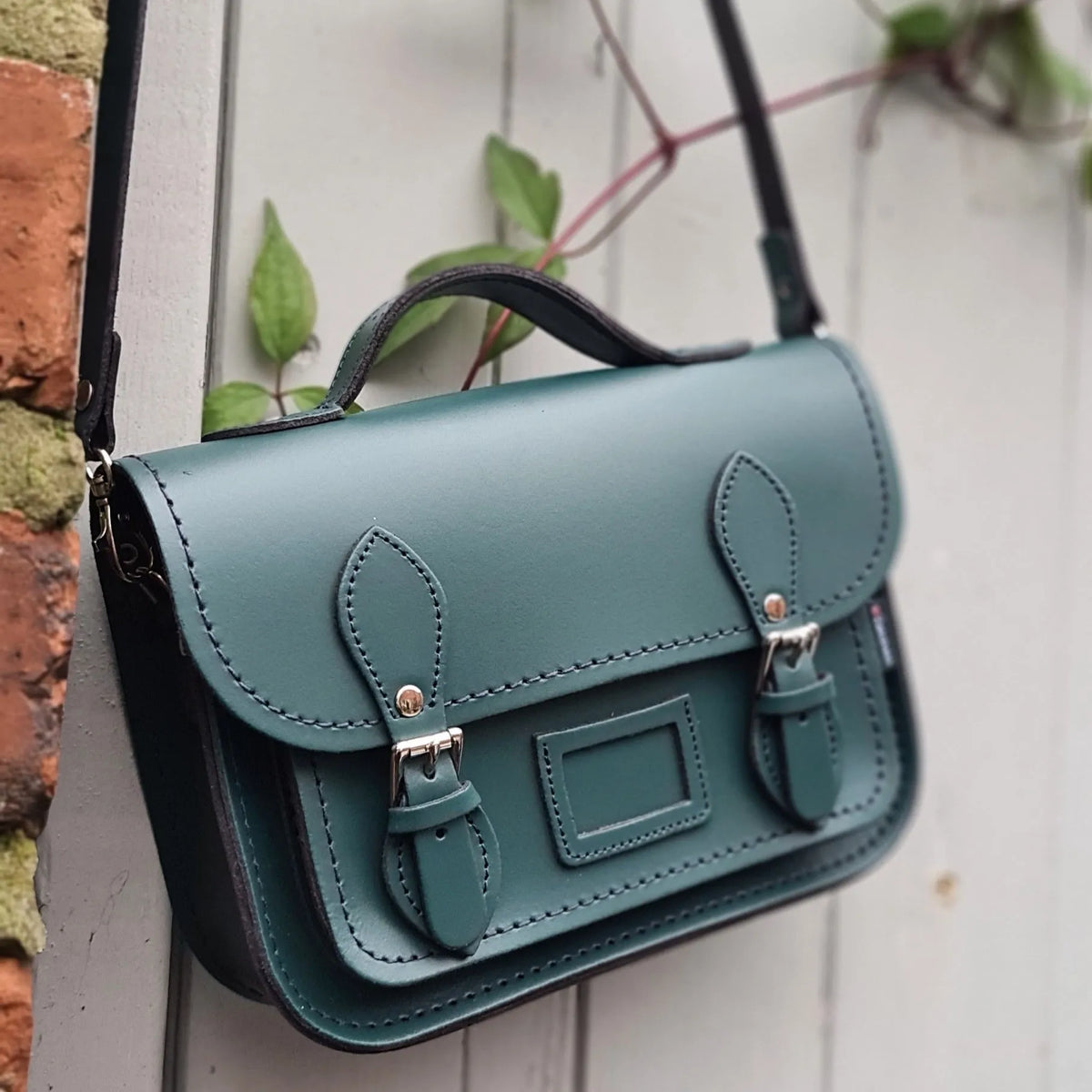 Zatchels Pine leather midi satchel hanging beside a brick wall and climbing leaves