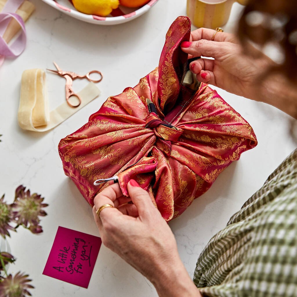 Person tying a red and gold fabric bundle on a white surface with scissors and flowers nearby.