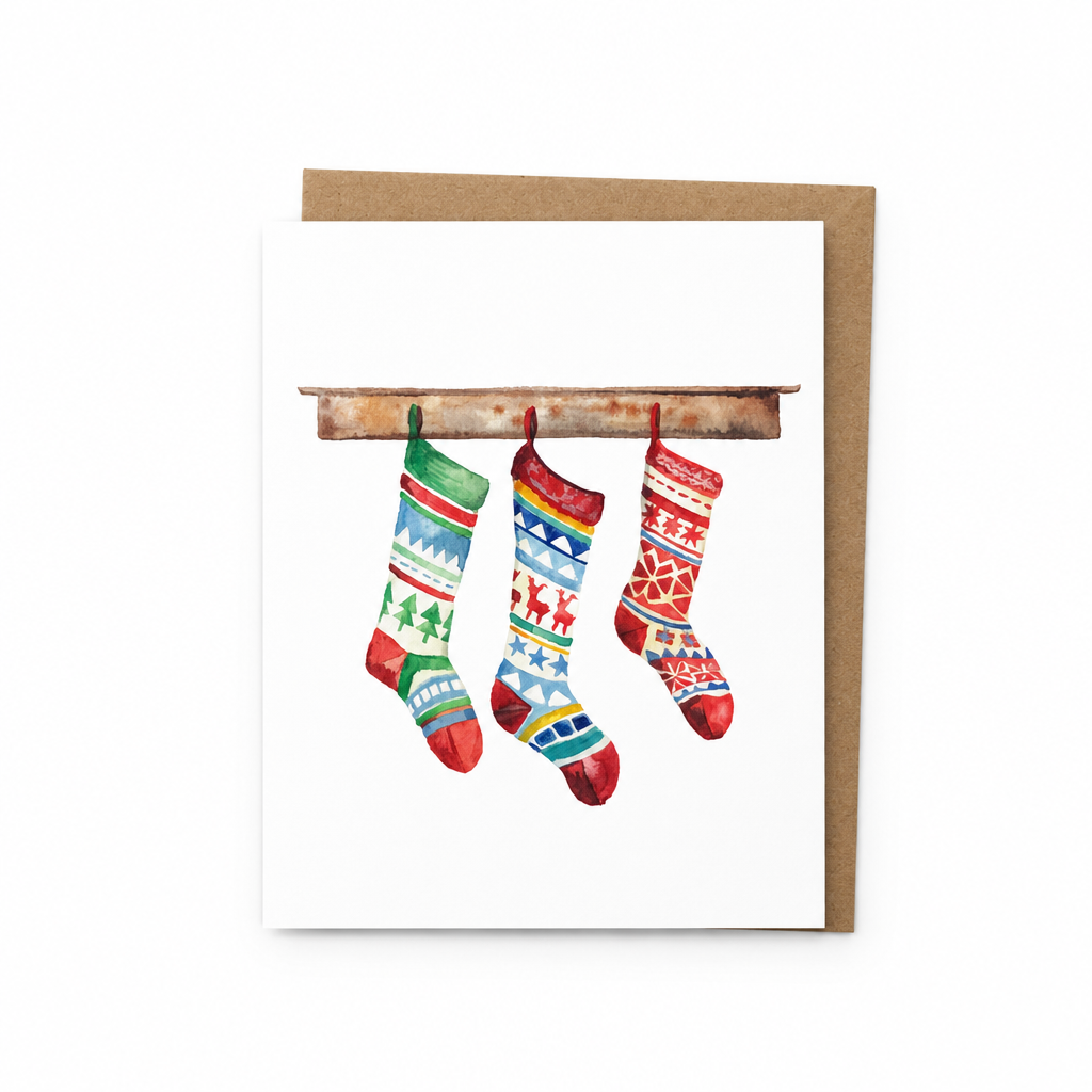 A white greeting card with a watercolor illustration of three colorful Christmas stockings hanging from a rustic wooden branch, shown with a brown kraft envelope behind it on a white background.