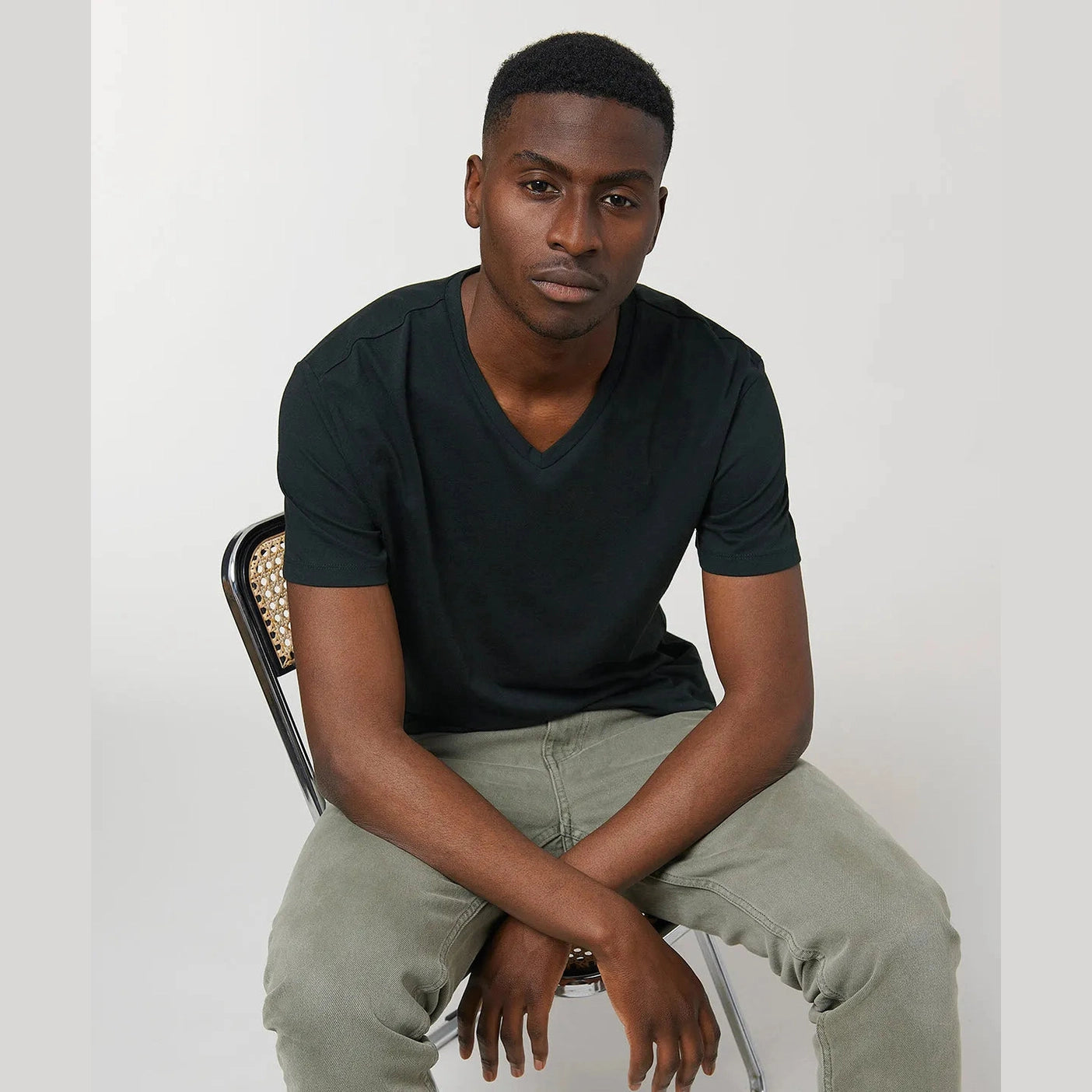 Man sitting on a chair wearing a dark green t-shirt and light gray pants against a plain background