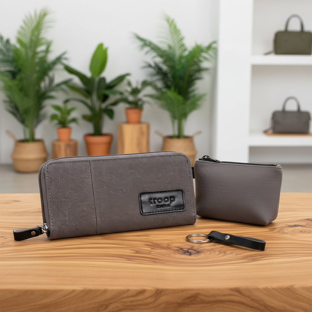 Two grey textured zippered pouches from Troop London on a wooden table, with a small black keychain tag in front. The background shows a blurred interior with potted plants and a shelf.